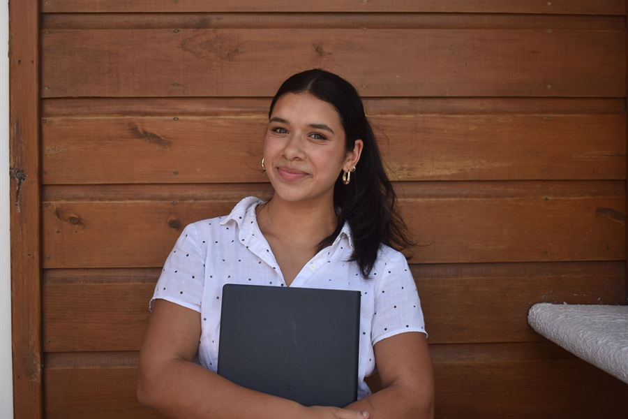 Sofia smiling warmly while holding her laptop in front of a wooden wall