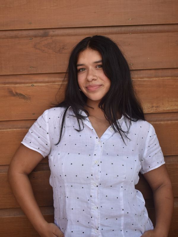 Sofia smiling in a white polka-dot shirt, hands in her pockets, in front of a warm wooden wall